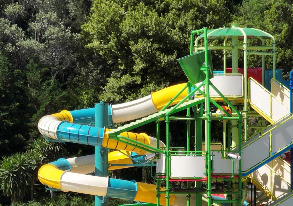 Ūdens slidkalniņš ar krāsainām caurulēm un struktūrām, kas atrodas zaļā apkārtnē. Ideāla vieta izklaidei un atpūtai ģimenēm.