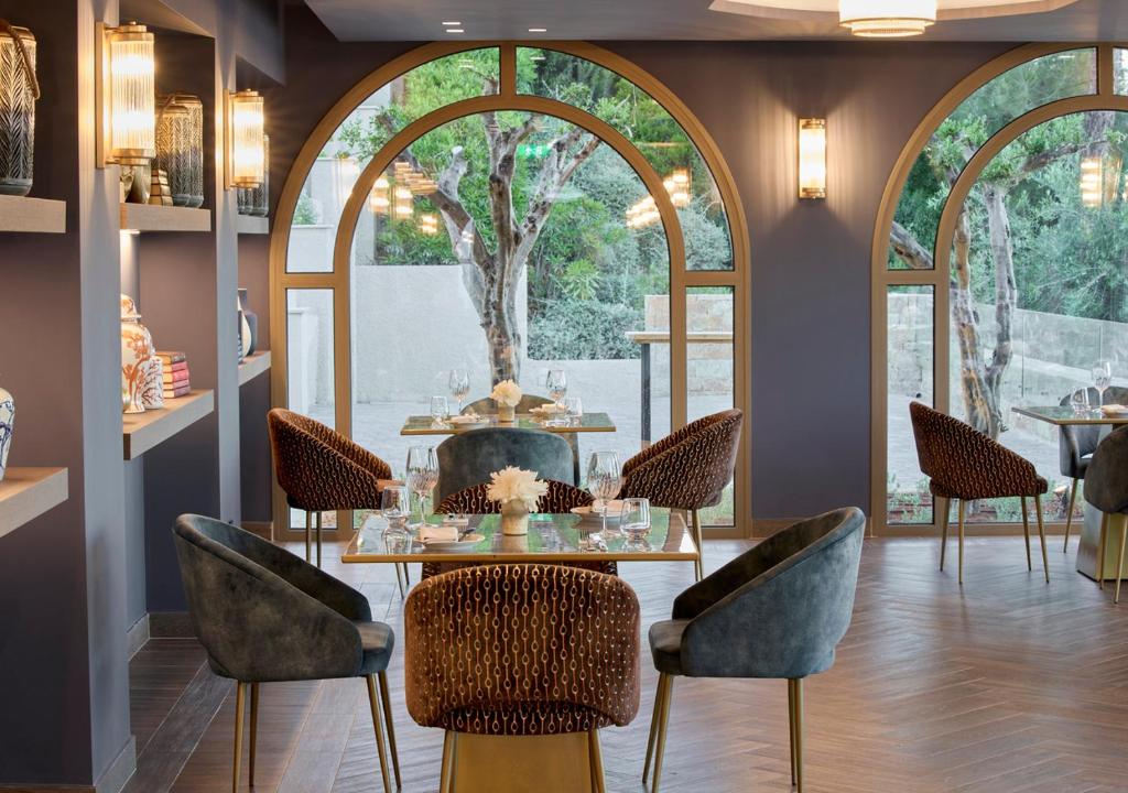 Mūsdienīga restorāna interjers ar elegantiem krēsliem, stikla logiem un dekoratīviem elementiem, kas rada patīkamu atmosfēru. Daba redzama caur logiem, piešķirot telpai dzīvīgumu.