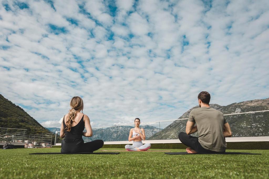 Trīs cilvēki meditē uz terases ar zaļu zāli, apkārt ir kalni un debesīs redzami mākoņi. Apskatāma mierīga un relaksējoša atmosfēra.