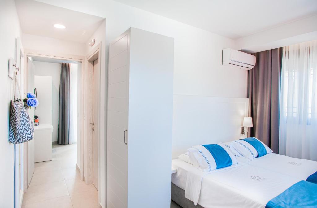 Mūsdienīga viesnīcas numura iekšskats ar gultu, baltiem un ziliem gultas piederumiem, gaismu un logu ar aizkariem. Interjers ir gaišs un elegants, radot patīkamu atmosfēru.