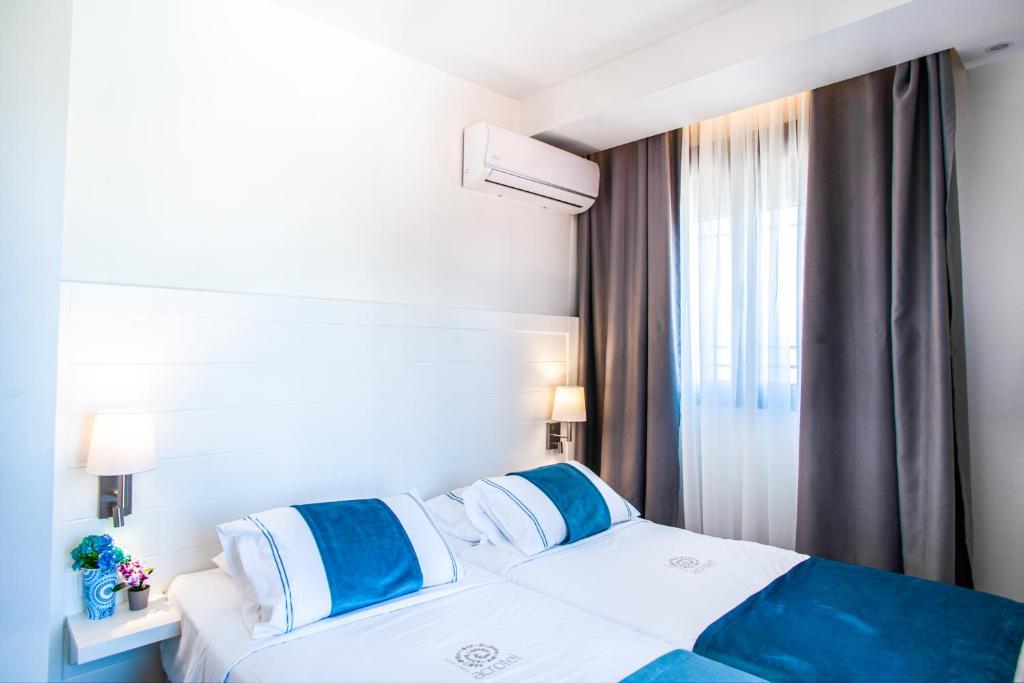 Mūsdienīgs guļamistabas interjers ar divām gultām, gaišām sienām un drēbēm zilā krāsā. Logā plūst dabiska gaisma, radot siltu atmosfēru.