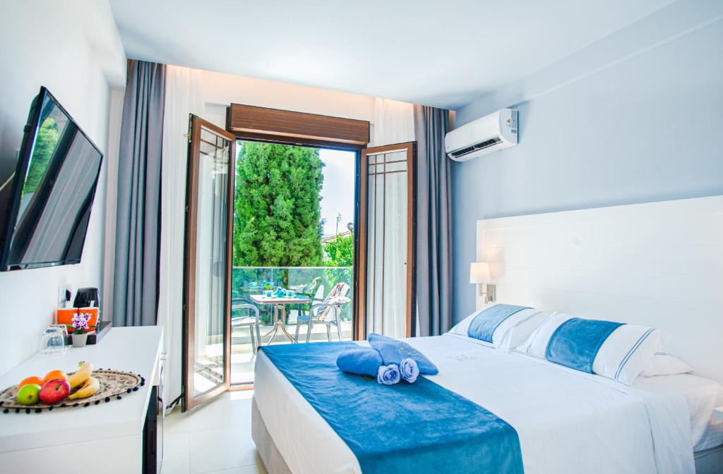 Mūsdienīga viesnīcas numura iekārtojums ar balkonā redzamu skatu uz zaļumiem. Gultā ir balts veļas komplekts ar zilām detaļām, un istabā ir televizors un mini ledusskapis.