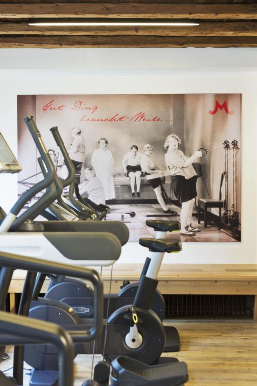 Sporta zāles interjers ar modernām trenažieru iekārtām un melnbaltu vintage fotogrāfiju uz sienas, kurā redzami cilvēki vingrojot.