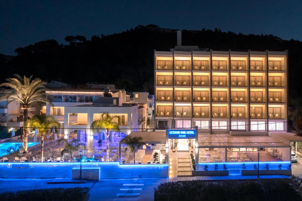Mūsdienīgs viesnīcas ēkas attēls naktī, apgaismots ar zilu gaismu, kas izceļ peldbaseinu un apkārtējos palmus. Viesnīcas nosaukums "Oceans Park Hotel" redzams ieejas daļā.