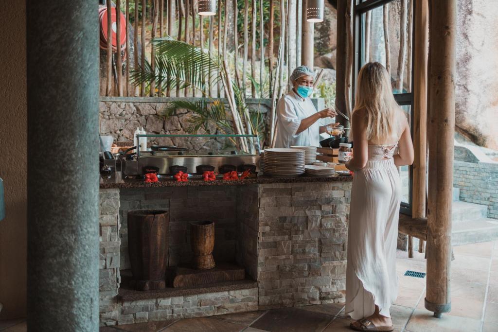 Bārs ar akmens sienām un koka elementiem, kur viesmīlis pasniedz ēdienu sievietei baltā kleitā. Apkārt augošas palmas un relaksējoša atmosfēra.