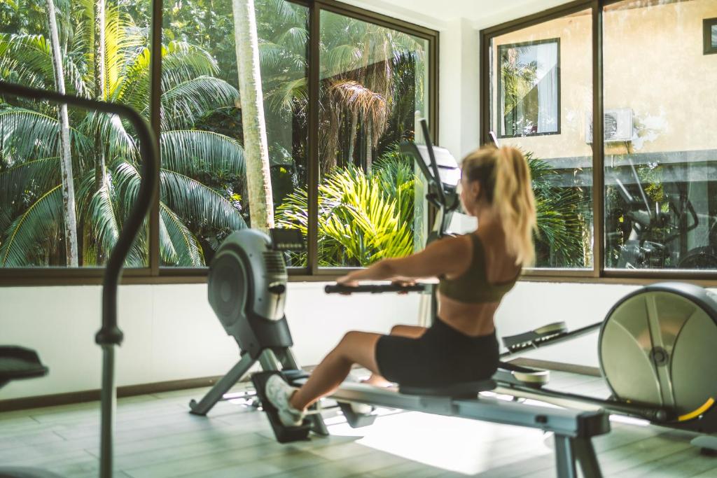 Sieviete sporto sporta zālē, izmantojot airēšanas mašīnu, kamēr caur logiem redzamas zaļas palmas.