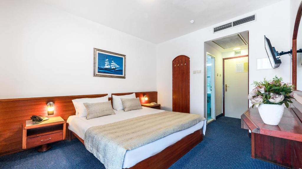 Mūsdienīgs viesnīcas numurs ar divguļamo gultu, koka mēbelēm un gleznu uz sienas, kas rada viesmīlīgu atmosfēru. Numurā ir televizors un neliels zieds uz galda.