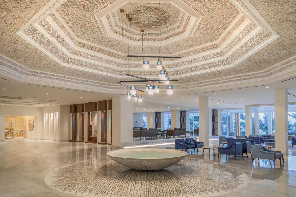 Elegants viesnīcas vestibils ar izsmalcinātu griestu dizainu un modernām mēbelēm. Centrā atrodas dekoratīvs ūdens elements, kas papildina kopējo atmosfēru.