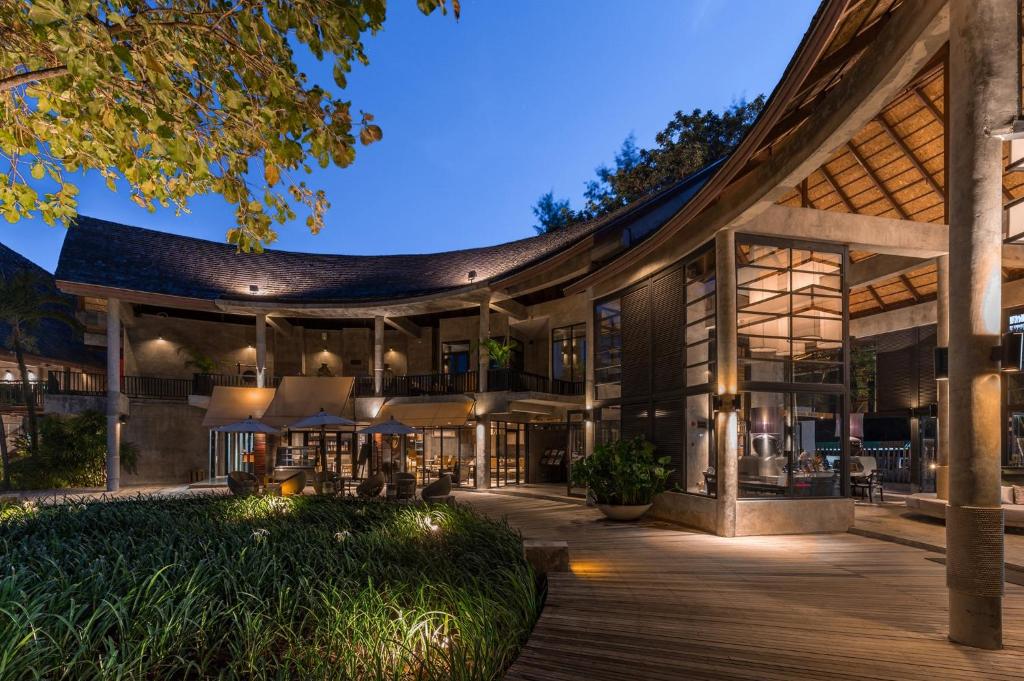 Mūsdienīga arhitektūra ar plašām stikla virsmām un koka elementiem, apgaismota naktī, radot siltu un viesmīlīgu atmosfēru. Apkārtne ir zaļa ar zālēm un augiem, kas papildina kopējo ainavu.