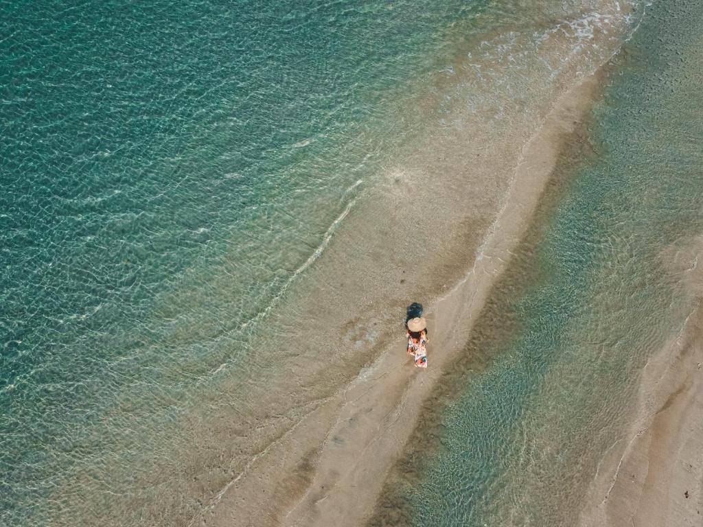 Kāds cilvēks staigā pa pludmali, kur ūdens ir dzidrs un sekls, radot mierīgu un saulainu atmosfēru.