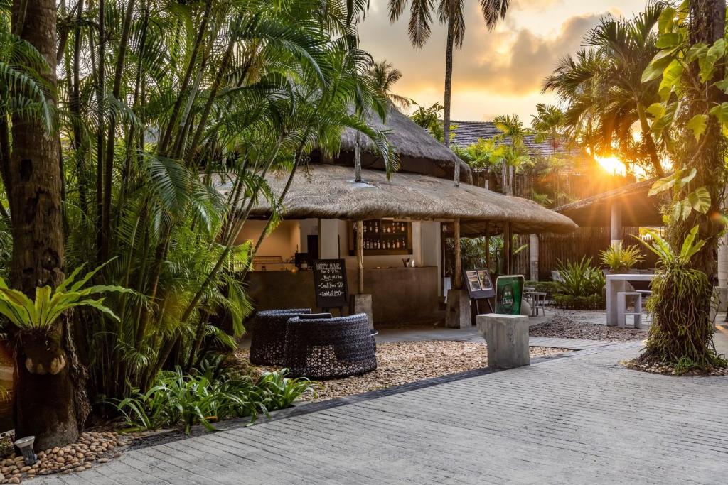 Tropu kūrorts ar džungļu ainavu, kur redzams bārs ar salmu jumtu un apkārtējiem augiem, saules stari izgaismo ainavu.