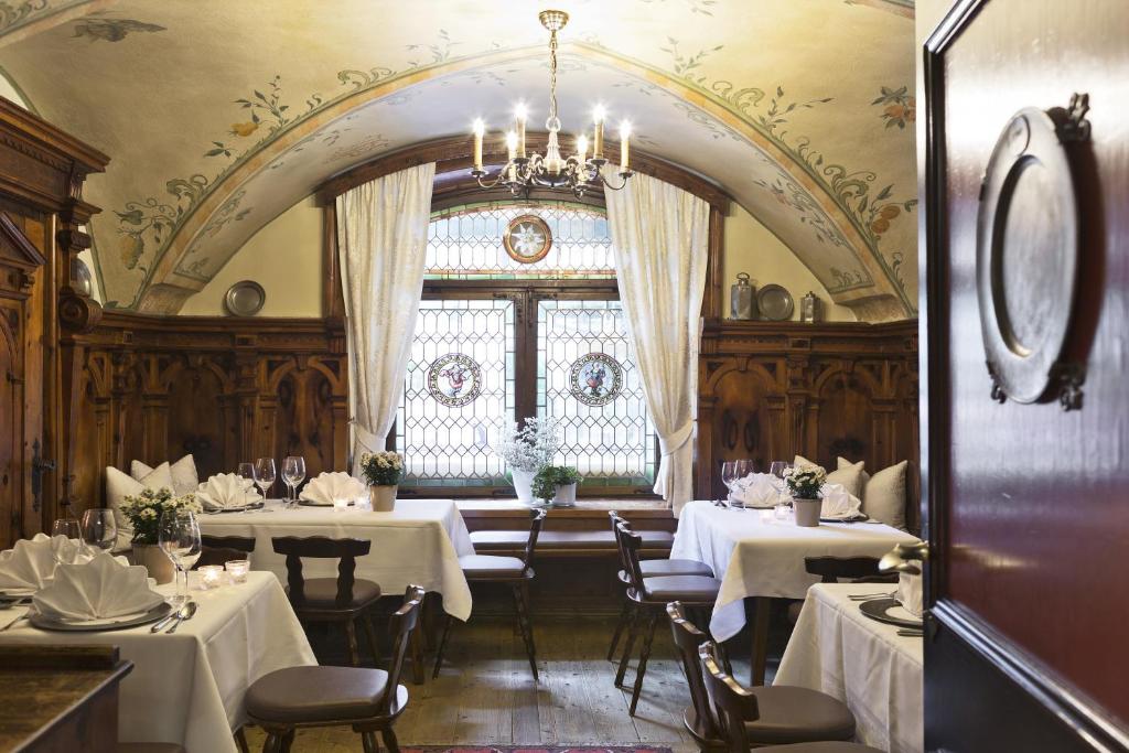 Eleganta restorāna iekārtojums ar koka apdari, baltiem galdautiem un ziedu dekorācijām, izgaismots ar elegantu lustru. Logi ar vitrāžām piedāvā gleznainu skatu uz ārpusi.