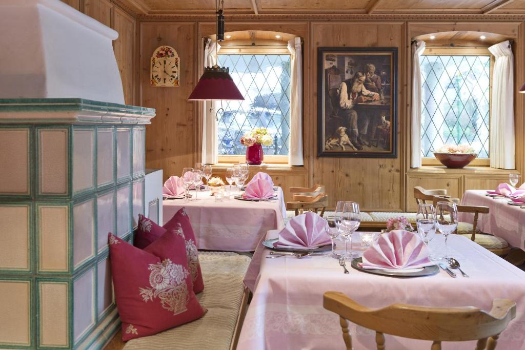 Skaista un eleganta restorāna iekārtojums ar rozā galda veļu, dekoratīviem spilveniem un mākslas darbu uz sienas. Gaismas logi un silta koka apdare rada mājīgu atmosfēru.