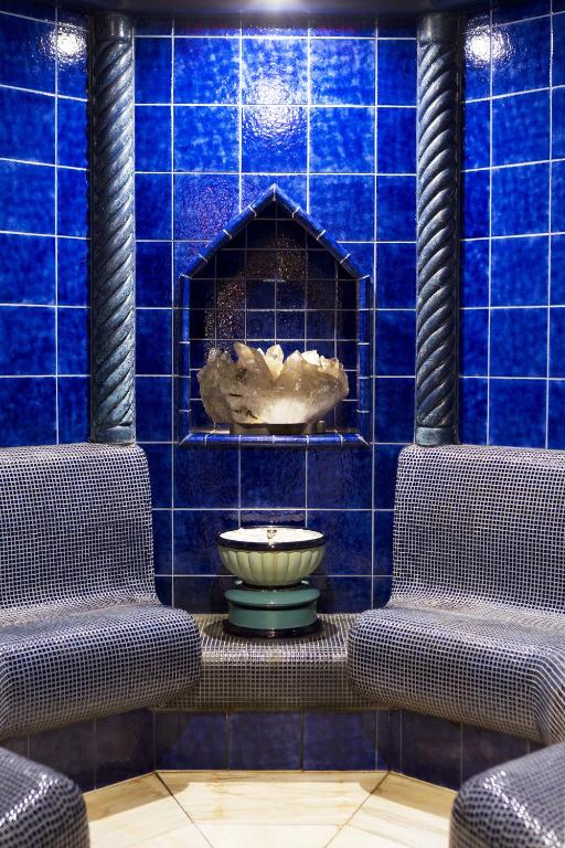 Mūsdienīgs relaksācijas stūris ar zilu flīžu sienām un divām ērtām sēdvietām, centrā atrodas dekoratīva ūdens strūklaka ar kristālu.