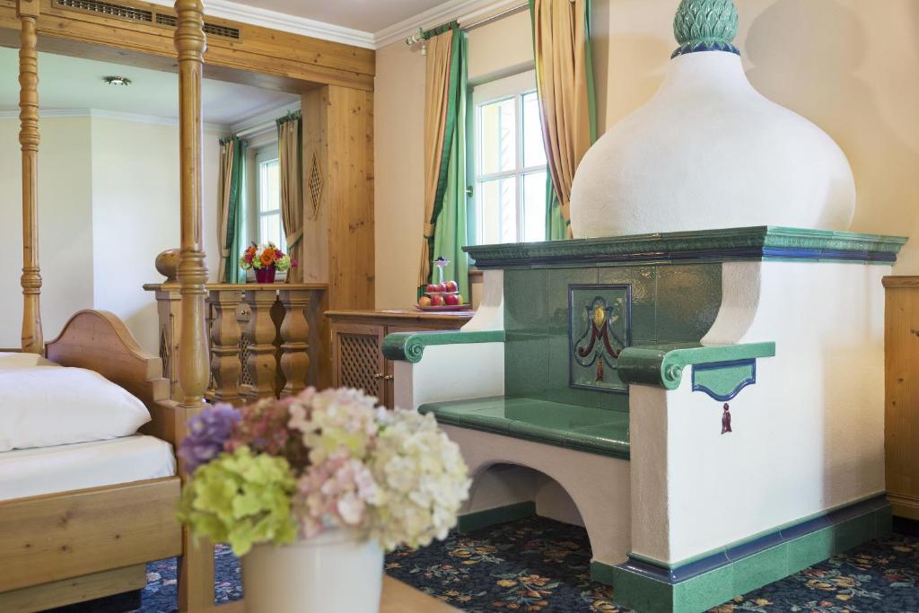 Mūsdienīga telpa ar gultu, koka mēbelēm un krāsainiem aizkariem, centrā izceļas stilizēts kamīns ar zaļām flīzēm. Uz galda novietoti ziedi un augļi, radot mājīgu atmosfēru.