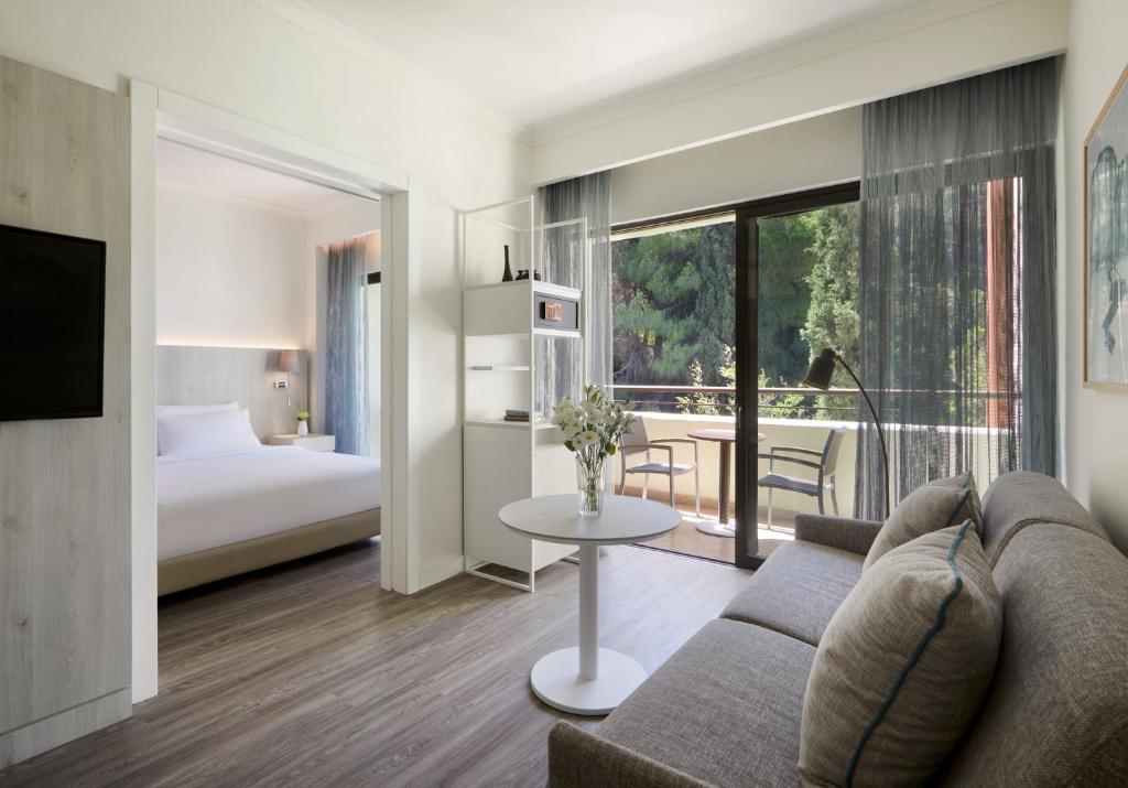 Mūsdienīgs viesnīcas numurs ar atsevišķu guļamistabu un dzīvojamo zonu, kas ietver dīvānu un galdu ar ziediem. Lieli logi nodrošina dabīgu apgaismojumu un skatu uz zaļo dabu.