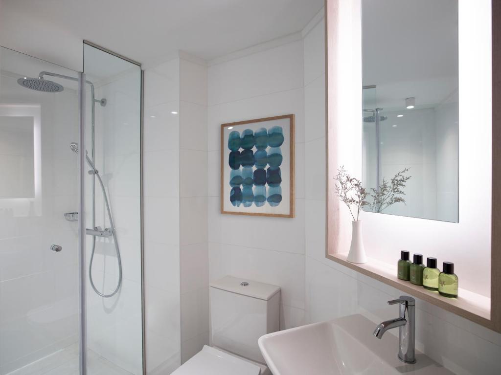 Mūsdienīgs vannas istabas interjers ar stikla dušu, baltiem flīzēm un dekoratīvu mākslas darbu uz sienas. Uz izlietnes novietoti zaļi kosmētikas produkti un ziedu kompozīcija.