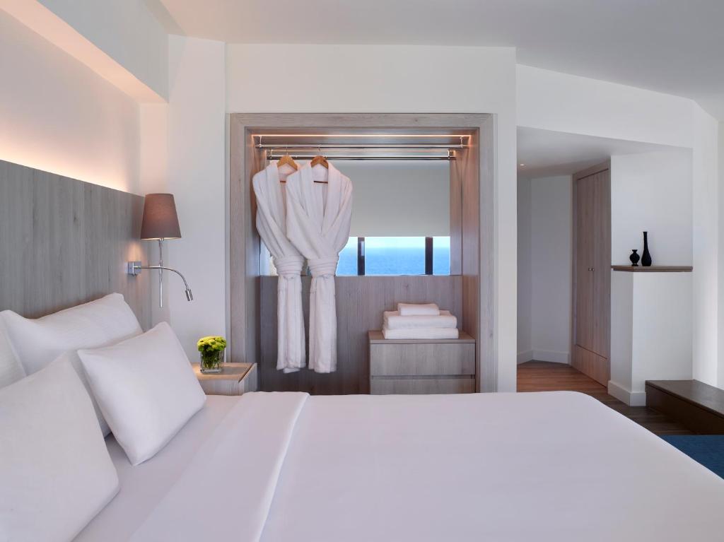 Mūsdienīgs viesnīcas numurs ar skatu uz jūru, kurā redzami balti gultas veļas komplekti un peldmēteļi, kā arī eleganta interjera elementi.