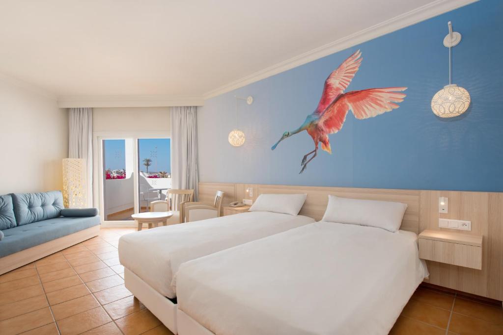 Mūsdienīgs viesnīcas numurs ar divām gultām, gaišām krāsām un māksliniecisku putna attēlu uz sienas. Telpā ir ērta dīvāns, koka mēbeles un daudz gaismas no logiem.