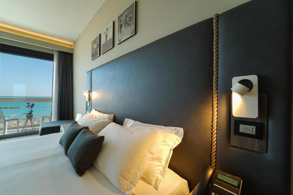 Mūsdienīgs viesnīcas numurs ar skatu uz jūru, kurā ir ērta gulta un eleganta apgaismojuma sistēma. Logā redzamas jūras viļņi un balkons ar ziediem.