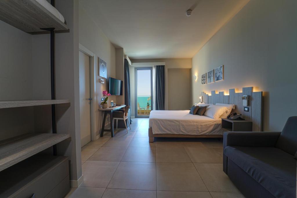 Mūsdienīgs viesnīcas numurs ar skatu uz jūru, kurā ir gulta, dīvāns un galds. Telpa ir gaiša un eleganta, ar neitrālām krāsām un minimālistisku dizainu.