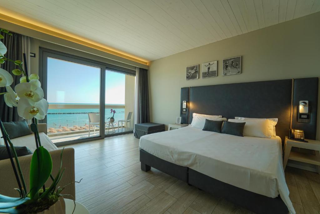 Mūsdienīgs viesnīcas numurs ar skatu uz jūru, kurā ir liela gulta, dīvāns un balkons. Interjers ir gaišs un elegants, ar orhidejām un mākslas darbiem uz sienām.