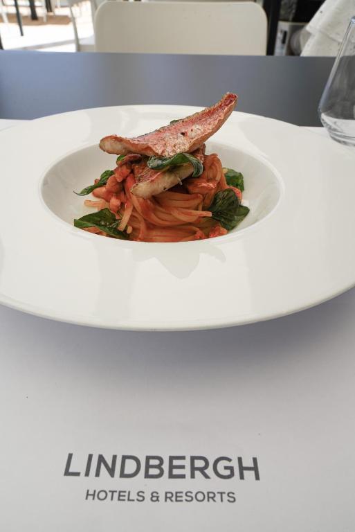 Mūsdienīgs ēdiens ar makaronu un zivi, pasniegts baltā traukā uz galdauta ar "Lindbergh Hotels & Resorts" logo.