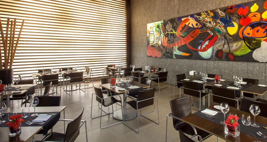 Mūsdienīgs restorāns ar elegantiem galdiem un krēsliem, ko izgaismo dabiska gaisma, un dekoratīvu mākslas darbu uz sienas. Stilīgas detaļas un sarkanas ziedu kompozīcijas piešķir atmosfērai īpašu šarmu.