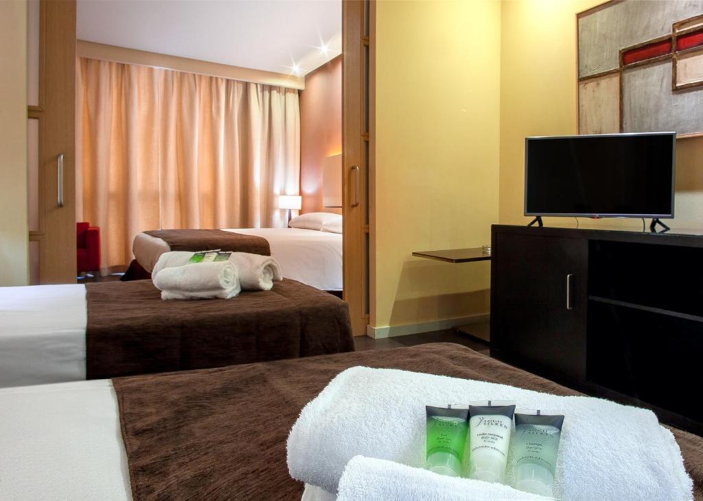 Moderns viesnīcas numurs ar divām gultām, televizoru un higiēnas līdzekļiem uz galda. Logi ar aizkariem nodrošina siltu un mājīgu atmosfēru.