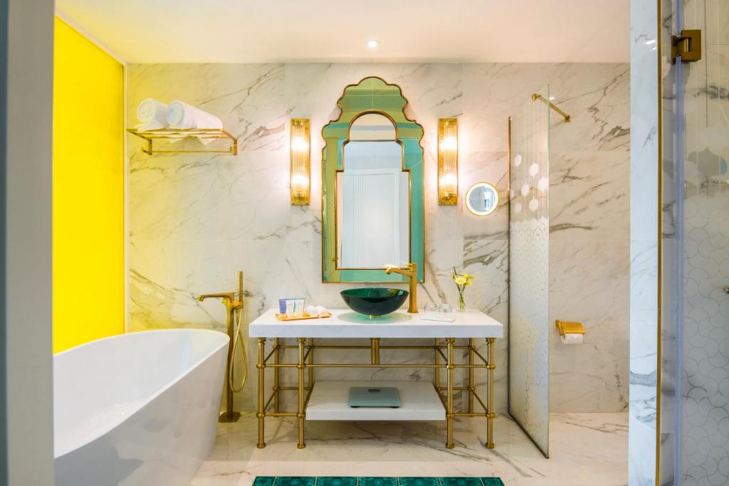 Mūsdienīgs vannas istabas interjers ar marmora sienām, zeltītām detaļām un spilgti dzeltenu akcentu, radot elegantu un stilīgu atmosfēru. Vannas istabā ir brīvi stāvoša vanna, moderns izlietnes galds un dekoratīvs spogulis.