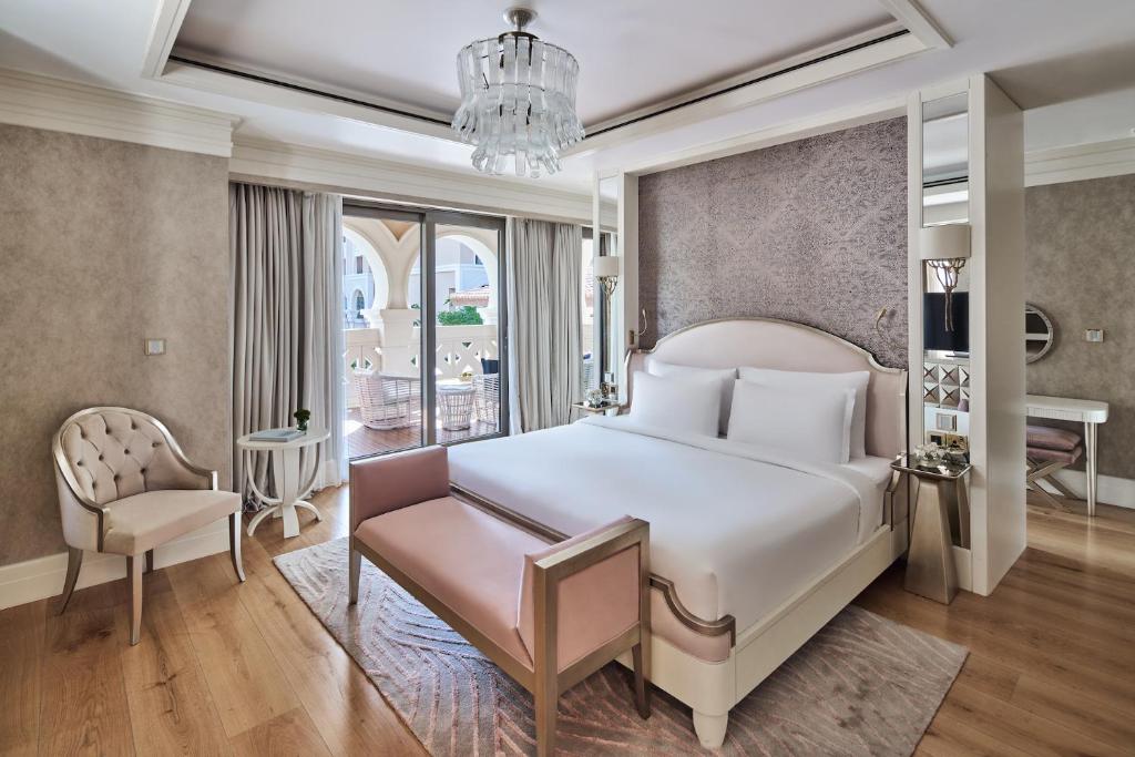 Elegantā viesnīcas numura interjers ar gultu, stilīgu krēslu un lieliem logiem, kas ved uz balkonu. Siltas krāsas un moderns dizains rada patīkamu atmosfēru.