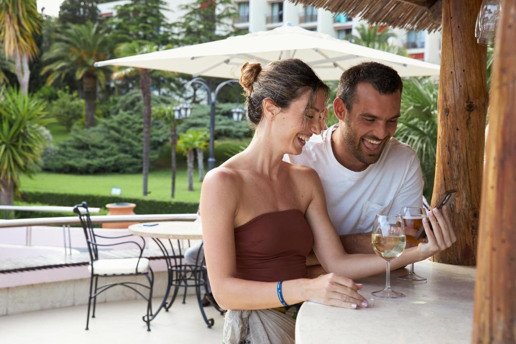 Jauns pāris izbauda brīvdienas, sēžot pie bāra un skatoties viedtālrunī, kamēr blakus ir divas dzērienu glāzes. Apkārt ir skaista dārza vide ar palmām.