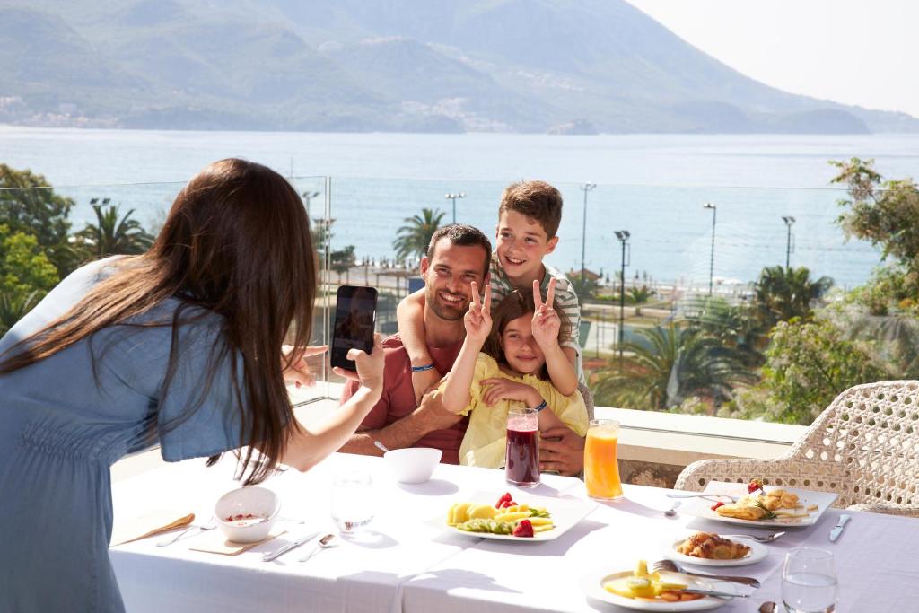 Ģimene, kas priecīgi pozē kopīgā fotoattēlā, sēžot pie galda ar ēdienu un dzērieniem, fona ir pludmale un kalni.