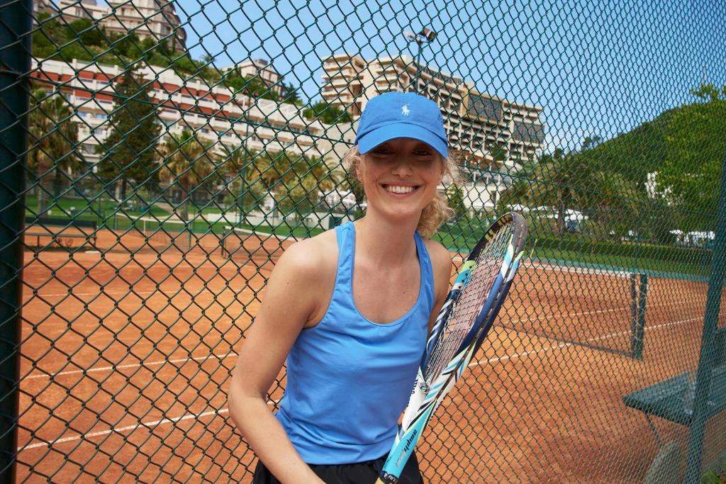 Smaidoša sieviete tenisa kortā, tērpta zilā tērpā un cepurē, tur tenisa raketi. Fons ietver zaļus kokus un sporta laukumus.