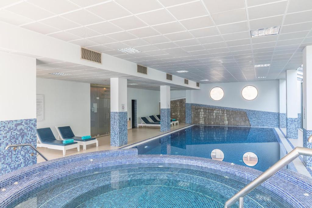 Moderns iekštelpu baseins ar relaksējošu burbuļu vannu, apgaismots ar dabīgo gaismu. Apkārt atrodas atpūtas krēsli un flīzētas sienas.