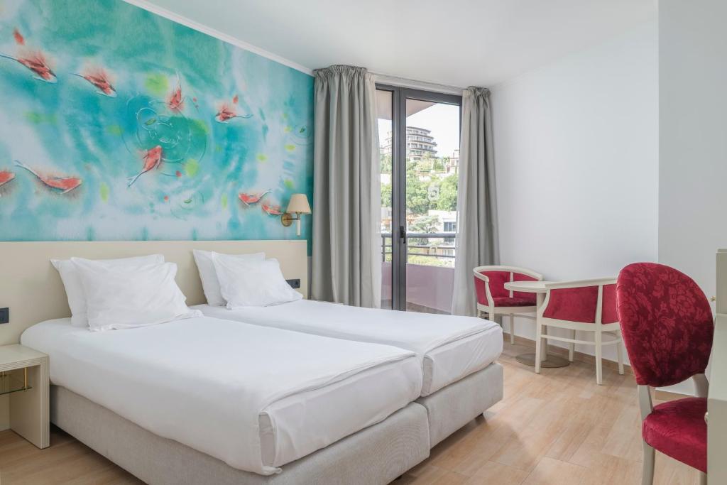 Mūsdienīgs viesnīcas numurs ar divām gultām, gaišām krāsām un māksliniecisku zīmējumu uz sienas. Numurā ir arī galds un krēsli, kā arī balkons ar skatu uz zaļo apkārtni.