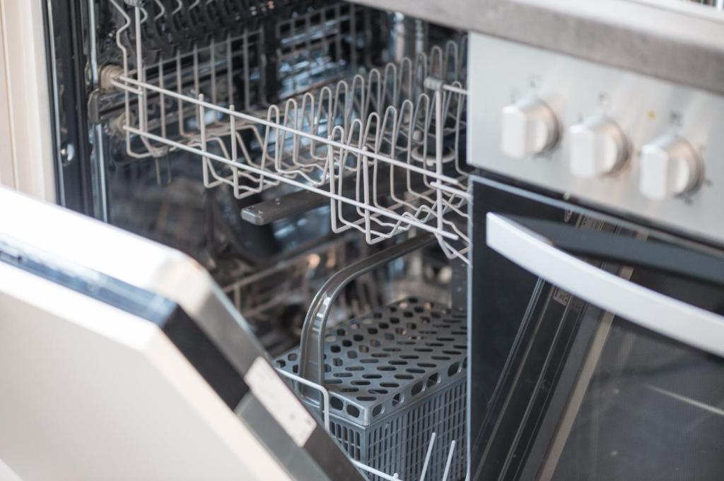 Aptiekts skats uz atvērtu trauku mazgājamo mašīnu, kurā redzami statīvi un grozi trauku novietošanai.