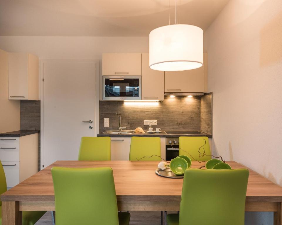 Modernā virtuve ar koka galdu un zaļām krēslām, gaiša apgaismojuma un minimālistiska dizaina elementi. Virtuvē ir iekļauts mikroviļņu krāsns un izlietne.