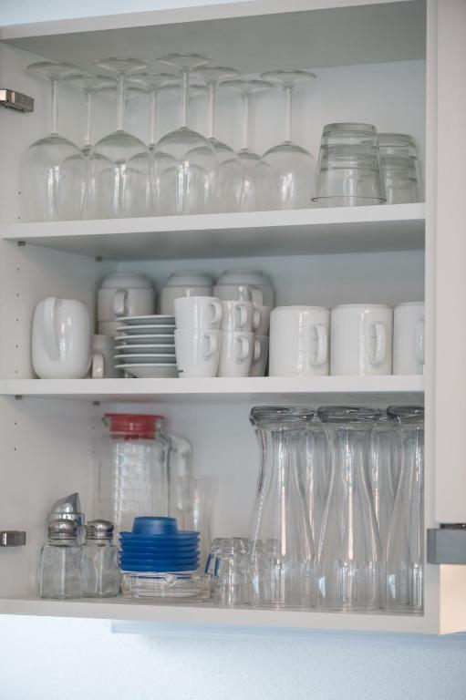Sakārtota virtuves skapja iekšpuse ar dažādiem traukiem, tostarp glāzēm, kafijas krūzēm un šķīvjiem. Trauki ir sakārtoti pa plauktiem, radot tīru un organizētu izskatu.