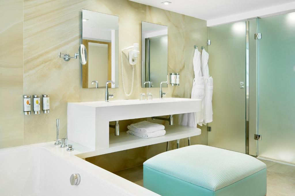 Mūsdienīgs un elegants vannas istabas interjers ar diviem spoguļiem, baltu vannas istabas mēbeli un stikla durvīm. Vannas istabā ir ērta pufīte un pieejami ķermeņa kopšanas līdzekļi.