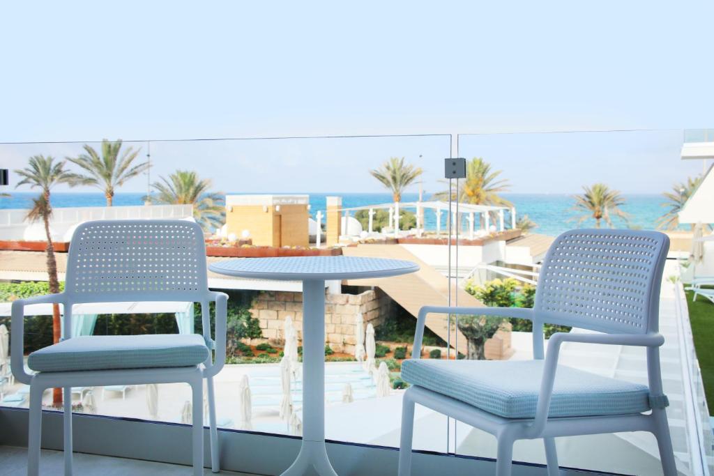 Mūsdienīgs balkons ar skatu uz jūru, kurā redzami divi baltas krēsli un apaļa galda. Fons veido zaļumi un palmām bagāta ainava.