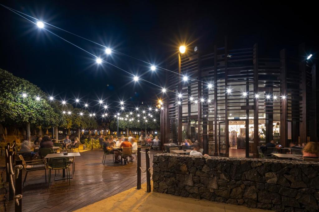 Skaista nakts ainava ar apgaismotu terasi, kur cilvēki bauda vakariņas zem zvaigžņotām debesīm. Mūsdienīga arhitektūra un zaļumi rada patīkamu atmosfēru.