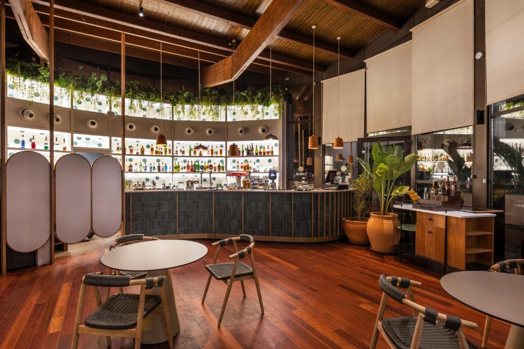 Mūsdienīgs bārs ar elegantu interjeru, kas ietver koka grīdu, dekoratīvas augu kompozīcijas un stilīgu bāra letes dizainu, aiz kura redzami dažādi dzērieni.