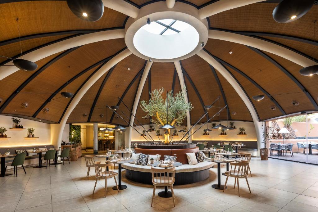 Mūsdienīgs restorāna interjers ar apaļu jumtu un elegantiem galdiem, kas rotāti ar augiem un dekoratīviem elementiem. Vieta ir gaiša un atvērtā, radot patīkamu atmosfēru.