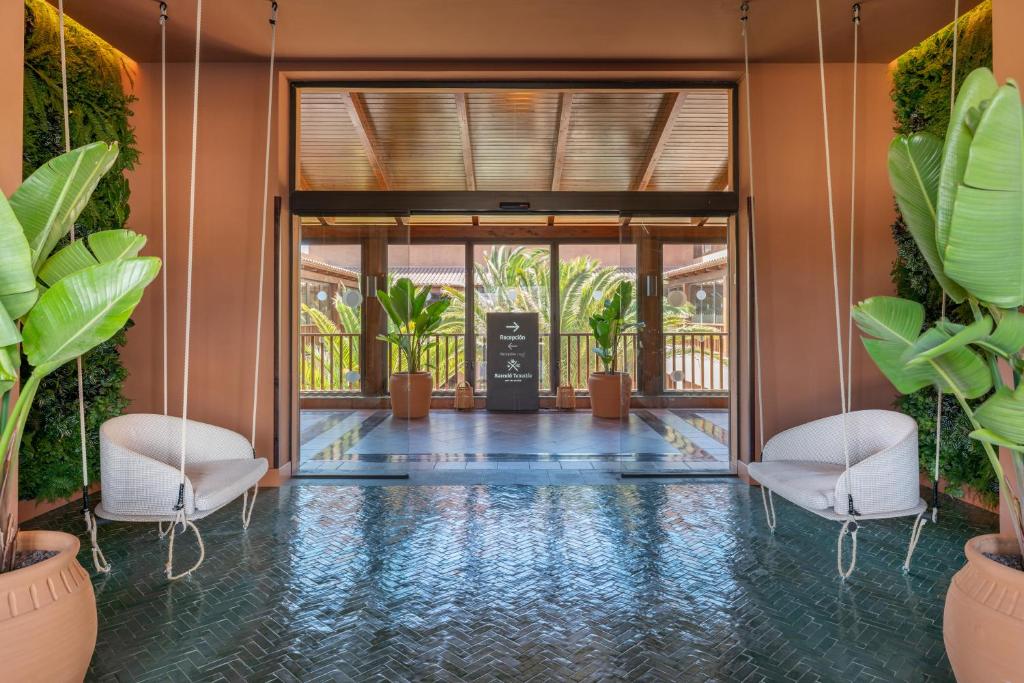 Mūsdienīgs viesnīcas interjers ar šūpuļkrēsliem un zaļām telpaugiem, radot relaksējošu atmosfēru. Plaša ieeja ar dabīgu apgaismojumu un elegantu dekoru.