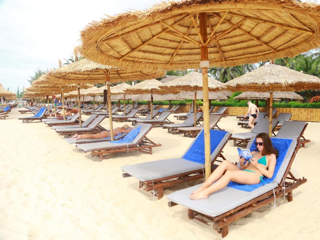 Pludmales atpūtas zona ar sauļošanās krēsliem un saules laidēm, kur viens cilvēks lasa grāmatu, bet citi atpūšas. Ideāla vide relaksācijai un atpūtai pie jūras.