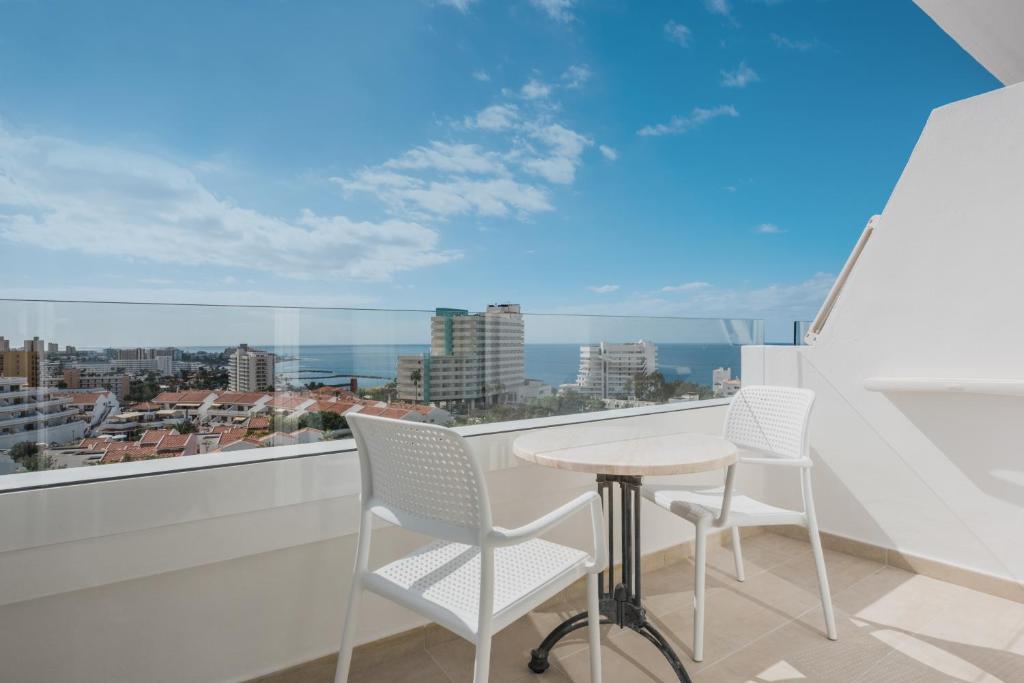 Balkons ar divām baltām krēslām un mazu galdiņu, skats uz pilsētu un jūru zem saulainām debesīm.