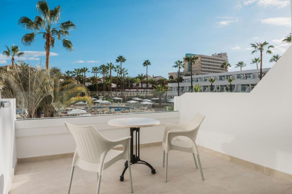 Balkons ar skatu uz palmām un viesnīcu kompleksu, kurā atrodas baltas krāsas krēsli un galds, radot relaksējošu atmosfēru.