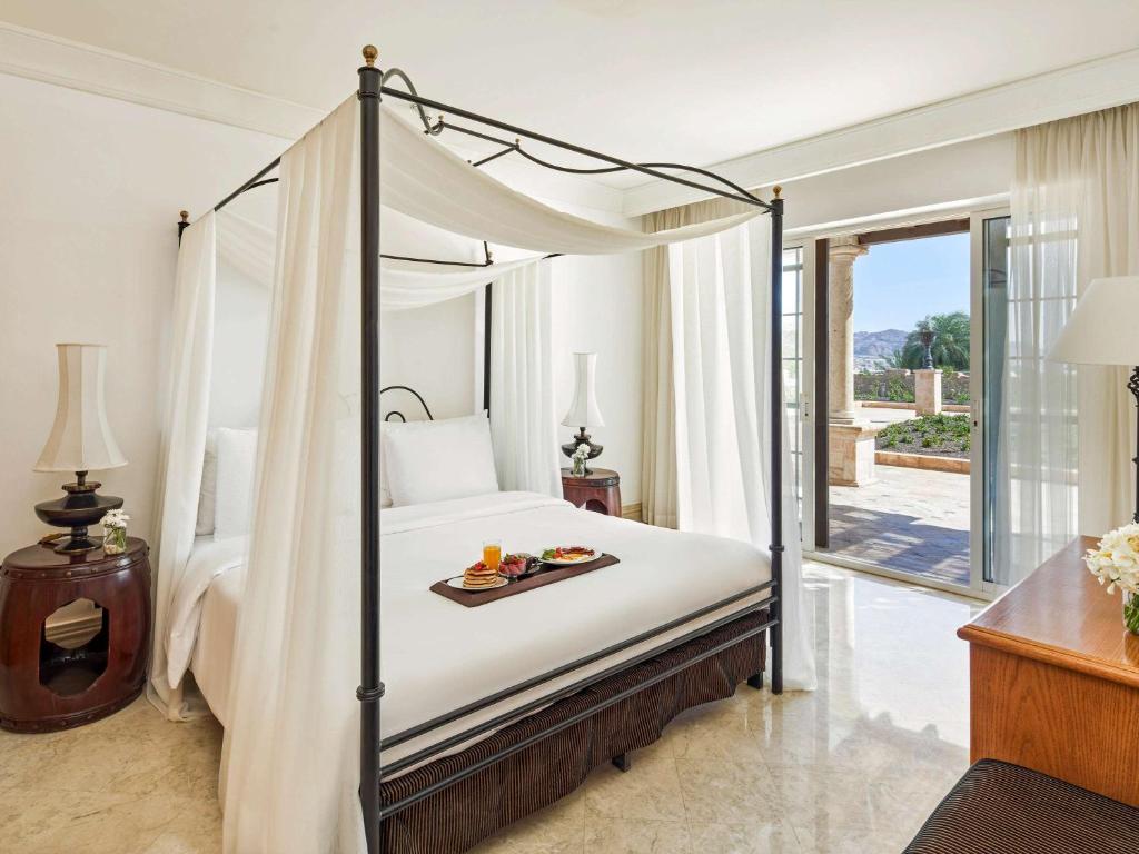 Eleganta guļamistaba ar četriem stabiem un baltiem aizkaru audumiem. Telpā ir moderna gulta, naktsgaldiņi un liels logs ar skatu uz dārzu.