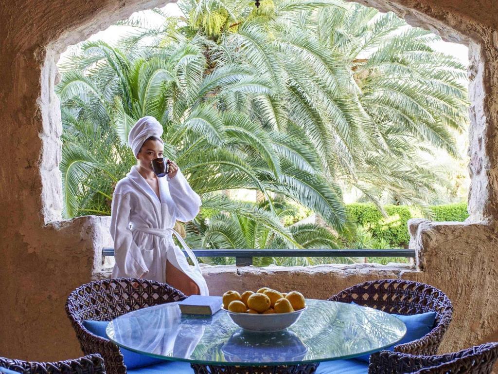 Sieviete baltā halātā un dvielī, turot tasi, stāv pie loga ar palmām fonā, blakus ir apaļa galds ar augļiem.